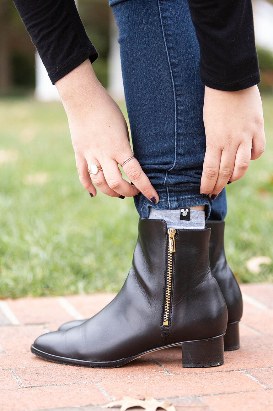 A pair of feet in black leather boots with a woman's hands pulling a jean leg up to show the zipper details